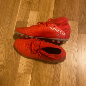 Röda Adidas Nemesis fotbollsskor - Säljer ett par röda Adidas Nemesis fotbollsskor med snörning och låg profil. Skorna har en cool randig textur och är designade för gräsplaner. Perfekta för dig som vill sticka ut på planen med stilrena och sportiga skor.