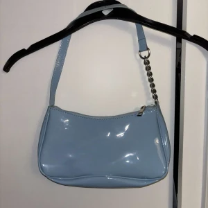Ljusblå axelväska med kedjedetalj - Snygg ljusblå axelväska i glansigt syntetmaterial med silverfärgad kedjedetalj på axelremmen. Väskan har dragkedja upptill och beige innerfoder. 