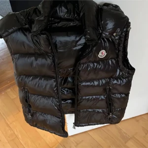 Svart dunväst från Moncler - Som ny.Säljer en svart, glansig dunväst från Moncler med hög krage och avtagbar huva. Västen har två fickor med dragkedja och klassisk Moncler-logga på bröstet. Insidan har en cool serietidningsdetalj. Perfekt för lager-på-lager och streetstyle.