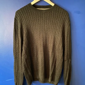 Mörkgrön stickad tröja från Crocker - Säljer en mörkgrön stickad tröja från Crocker i storlek M. Tröjan har ett klassiskt kabelstickat mönster, rund halsringning och långa ärmar. Perfekt för dig som gillar en stilren och tidlös look med lite extra struktur.