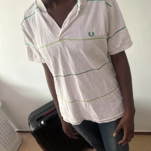 Vit randig t-shirt från Fred Perry - Vit t-shirt från Fred Perry med tunna gröna och gula ränder. Klassisk pikékrage och korta ärmar. Broderad logga på bröstet. Perfekt för en chill och sportig look.