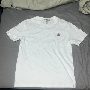 Vit Moncler t-shirt med logga - Vit t-shirt från Moncler i mjuk bomull med klassisk passform. T-shirten har rund halsringning och en broderad Moncler-logga på bröstet. Perfekt basic-plagg med exklusiv känsla och stilren look.