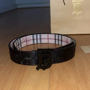 Svart bälte  - Snyggt svart bälte i skinn med klassiskt rutigt mönster på insidan och stort svart monogramspänne i metall. Bältet har bred design som ger en lyxig känsla. Perfekt accessoar för att lyfta din outfit.