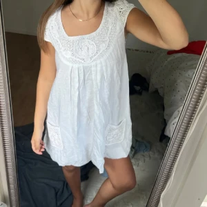 Vit kort klänning med spetsdetaljer - Supersöt vit kortklänning med spetsdetaljer vid halsringningen och på fickorna. Klänningen har korta ärmar och en lös, luftig passform som är perfekt för varma dagar. Materialet känns lätt och bekvämt, och de broderade detaljerna ger en romantisk vibe.