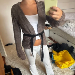 Brun stickad kofta från Stockhlm - Säljer en brun stickad kofta från Stockhlm i storlek XS. Koftan har ett snyggt kabelstickat mönster, långa ärmar och ett svart knytband i midjan. Perfekt för lager-på-lager och chill höst- eller vinterstil.