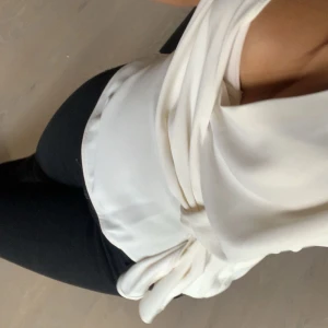 Vit omlott blus med knyt - Säljer en stilren vit omlott blus med snygg knytning i midjan. Blusen har en djup v-ringning och är ärmlös, vilket ger en fräsch och modern look. Perfekt att matcha med svarta byxor för en clean outfit.