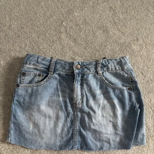 Blå jeanskjol med fickor - Säljer en klassisk blå jeanskjol i kort modell med slitna detaljer. Kjolen har fram- och bakfickor, bälteshällor och knappstängning framtill. Perfekt för dig som gillar en avslappnad och trendig stil.