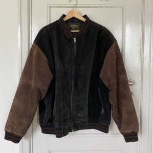 äkta mockajacka från cambridge classics - brun-svart collegejacka i äkta mocka från cambridge classics med randiga detaljer. jacakan har svarta och bruna paneler med ribbade muddar och krage i lila och brunt, dragkedja framtill och klassisk bomberpassform. perfekt för retrovibbar till hösten!