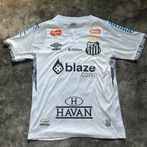 Santos neymar tröja - Santos FC matchtröja i vitt från Umbro med Neymar Jr och nummer 10 på ryggen. Tröjan har korta ärmar, svarta detaljer och flera tryckta sponsorer. Tillverkad i lätt och ventilerande material, perfekt för fotbollsälskare och samlare. Som ny!