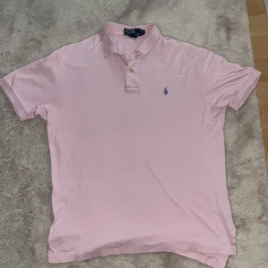 Rosa pikétröja från Polo Ralph Lauren - Säljer en klassisk rosa pikétröja från Polo Ralph Lauren i 100% bomull. Tröjan har korta ärmar, krage och knäppning framtill samt den ikoniska blå loggan broderad på bröstet. Perfekt för en clean och stilren look. Storleken är S men passar också M.