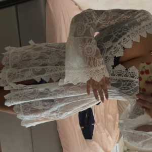 Vit spets coverup med knytband - Superfin kort vit spets bikini coverup med långa ärmar och knytband i midjan. Coverupen har vackra broderade detaljer och genombrutet spetsmönster som ger en romantisk vibe. Perfekt för sommarens alla tillfällen!