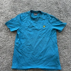 Blå t-shirt från Lyle & Scott - Snygg blå t-shirt från Lyle & Scott med klassisk gul logga broderad på bröstet. T-shirten har rund halsringning och korta ärmar. Perfekt basic-plagg med skön passform i mjuk bomull.