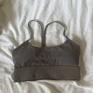 Grå sporttopp från ICANIWILL - Sporttopp i grå nyans från ICANIWILL med smala axelband och brottarrygg. Snygg minimalistisk design med logga framtill. Tillverkad i stretchigt syntetmaterial som passar perfekt för träning och aktiva dagar.