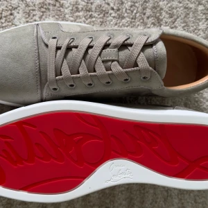 Grå mockasneakers från Louboutin - Snygga sneakers i beige mocka från Christian Louboutin med ikonisk röd sula. Skorna har snörning och en stilren, rund tå. Insidan är i lyxigt skinn och utsidan i mjuk mocka. Perfekta för dig som vill sticka ut med exklusiv känsla.