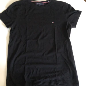 Svart t-shirt från Tommy Hilfiger - Klassisk svart t-shirt från Tommy Hilfiger i bomull. Diskret logga broderad på bröstet och rund halsringning. Enkel och stilren modell med korta ärmar, perfekt till jeans eller shorts.