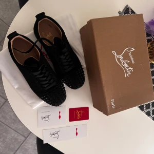 Christian Louboutin svarta sneakers med nitar - Svarta sneakers från Christian Louboutin i mocka med coola svarta nitar på tån och klassisk röd sula. Skorna har snörning och rund tå, vilket ger en edgy och lyxig vibe. Perfekta för dig som vill sticka ut med exklusiva detaljer.