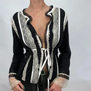 Svart spetsblus med snörning - Supersnygg svart blus med långa ärmar, dekorerad med vita spetsdetaljer och volangkant. Djup v-ringning och knytband framtill ger en unik look. Transparenta spetsinslag vid ärmslut och längs hela framsidan. Perfekt för dig som gillar statement-plagg.