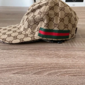 Gucci keps - Exklusiv keps från Gucci i beige och brunt GG-monogram över hela kepsen. Klassisk böjd skärm och ikoniska gröna och röda ränder på sidan. Tillverkad i bomullscanvas med brunt innerfoder och märkning på insidan.