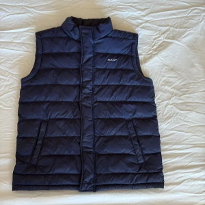 Mörkblå dunväst från Gant - Snygg mörkblå dunväst från Gant med quiltad design och hög krage. Västen har två fickor framtill och en insida i starkt röd färg. Perfekt för lager-på-lager och ger en sportig vibe till din outfit.