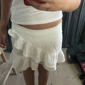 Vit volangkjol från Gina Tricot - Supersöt vit kortkjol från Gina Tricot med volangkant och elastisk midja. Kjolen är i ett lätt och luftigt bomullstyg.