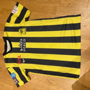 Al-Ittihad Benzema matchtröja Nike - Gul och svart randig Al-Ittihad fotbollströja med Benzema och nummer 9 på ryggen. Tröjan har korta ärmar, flera sponsorloggor och klubbmärke på bröstet. Tillverkad i lätt och ventilerande material, perfekt för fotbollsplanen eller att bära som supporter.