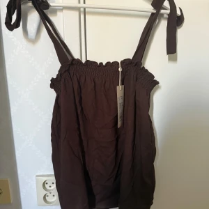 Brun croppad linnetopp från Cream - Snygg brun croppad linnetopp från Cream med smock upptill och knytbara axelband. Perfekt för varma dagar och enkel att styla med jeans eller kjol. Luftig och skön känsla tack vare linnematerialet.