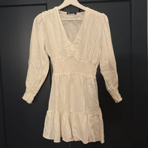 Krämvit kortklänning från Zara - Supersöt krämvit kortklänning från Zara med v-ringning, smockad midja och långa ärmar med smock vid ärmslut. Klänningen har volang nertill och är perfekt för en fräsch och trendig look. Lätt och luftig, passar perfekt till varmare dagar.
