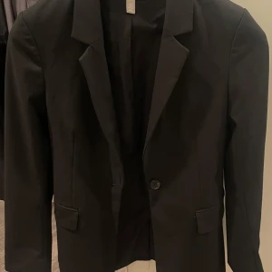Svart kavaj från H&M - Klassisk svart kavaj från H&M med enkelknäppning och snygg krage. Rak passform och diskreta fickor framtill. Perfekt att styla med skjorta eller t-shirt för en clean look.