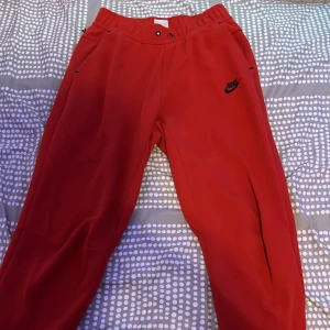 Röda mjukisbyxor från Nike - Röda mjukisbyxor från Nike med svart logga på ena benet. Byxorna har elastisk midja, sidfickor och en bakficka med dragkedja. Perfekta för chill eller träning, tillverkade i mjukt material som känns skönt mot huden.