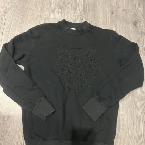 Svart Kenzo sweatshirt med tigerbrodyr - Svart sweatshirt från Kenzo med ikonisk tigerbrodyr och texten 'Kenzo Paris' framtill. Tröjan har rund hals, långa ärmar och ribbade muddar. Perfekt för dig som gillar streetstyle och vill sticka ut med en klassisk designertröja.