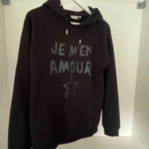 Gina Young hoodie - Svart hoodie med texten 'JE M'EN AMOUR' och en stjärna tryckt på framsidan. Tröjan har huva med snörning och långa ärmar. Perfekt för en avslappnad och cool stil.