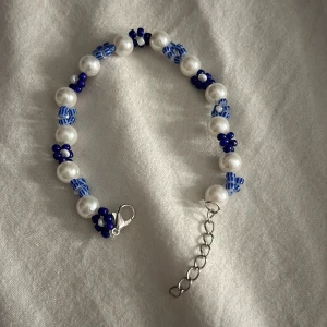 Pärlarmband med blåa detaljer - Superfint armband med vita pärlor och blåa glaspärlor i blommönster. Armbandet har silverfärgat spänne och justerbar kedja. Perfekt accessoar för att ge din outfit en söt och trendig touch. Gjort själv 