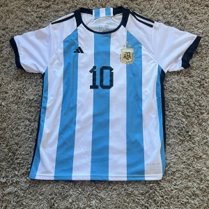Argentina Messi #10 Adidas fotbollströja - Säljer en  Argentina landslagströja från Adidas med Messi och nummer 10 på ryggen. Tröjan är kortärmad, har klassiska blå och vita ränder, broderat AFA-märke med tre stjärnor och Adidas-logga. Tillverkad i lätt och ventilerande polyester. Använd en gång