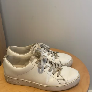 Vita sneakers från Michael Kors - Snygga vita sneakers från Michael Kors med beige detaljer och guldig MK-loggor på snörningen och hälen. Leather upper och tygdetaljer i beige med diskret logomönster. Perfekta för en clean och trendig look. Storlek 37 men är stora i storleken så passar 38 