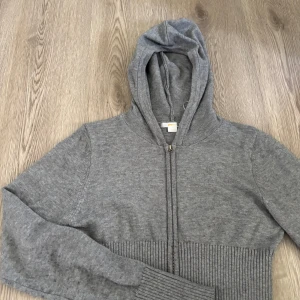 Grå stickad hoodie från Gina Tricot - Mysig grå hoodie från Gina Tricot i stickat material. Tröjan har dragkedja framtill, huva och breda ribbade muddar vid midja och ärmslut. Den är lite nopprig på armarna.