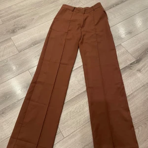 Bruna kostymbyxor med raka ben - Snygga bruna kostymbyxor med pressveck och raka ben. Byxorna har midjehällor och sidfickor, perfekta för en clean och stilren look. Materialet känns mjukt och har en lätt glansig finish.