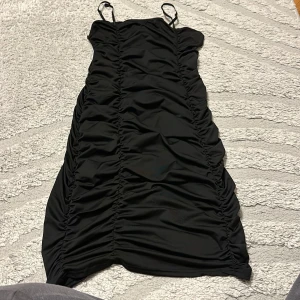 Svart ruched kortklänning - Snygg svart kortklänning med smala axelband och ruched detaljer längs hela klänningen. Figurnära passform som framhäver siluetten och ger en trendig look. Perfekt för dig som vill sticka ut med en stilren och modern vibe.