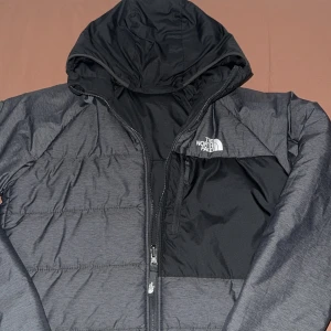 Svart pufferjacka från The North Face - Snygg svart pufferjacka från The North Face med huva och dragkedja framtill. Jackan har en stilren design med logga på bröstet och en praktisk bröstficka med dragkedja. Perfekt för kalla dagar och enkel att matcha med allt. Kan användas till båda håll