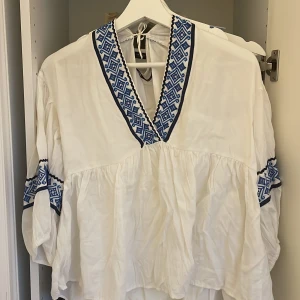 Vit och blå, sommarblus från Zara - Superfin vit blis från Zara med blå broderade detaljer och v-ringning. Trekvartsärm som är lite puffig. Perfekt för sommarens alla tillfällen, tillverkad i mjuk bomull. Snyggt mönster på ärmar och halsringning. 