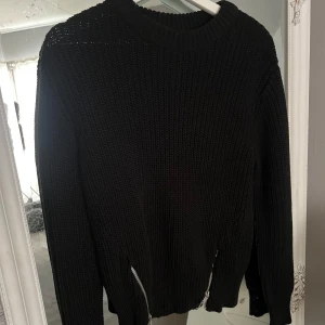 Svart stickad tröja med dragkedjor - Snygg svart stickad tröja med grov struktur och coola silverfärgade dragkedjor nertill på sidorna. Tröjan har rund halsringning och långa ärmar, perfekt för lager på lager under kyliga dagar.