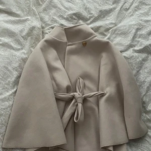 Beige kappa med bälte och vida ärmar - Säljer en superfin beige kappa med hög krage, knytbälte i midjan och vida ärmar. Kappan har en snygg guldig knapp upptill och är i ett mjukt ull-liknande material. Perfekt för dig som vill ha en stilren och trendig look.