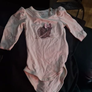 Söt naturvit body med igelkotten med rosett - Naturvit långärmad body från Name it i ekologisk bomull. Framsidan har ett gulligt tryck av en igelkotte med en liten rosett i tyg och glittriga detaljer. Fina puffärmar och spetskant vid halsen. Perfekt för de allra minsta.