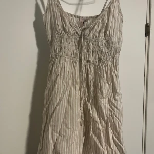 Randig sommarklänning från Nelly XS - Supersöt kortklänning från Nelly i ljusa beige och vita ränder. Klänningen har smala axelband, smockad midja och är tillverkad i ett lätt och luftigt material. Perfekt för varma dagar och ger en fräsch och avslappnad look.