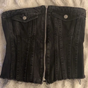 Svart jeanskorsett från Zara - Cool svart korsettopp i jeans från Zara med dragkedja framtill och råa kanter nertill. Toppen har dekorativa sömmar, fickdetaljer med knappar och smockad baksida för extra passform. Perfekt för dig som vill ha en edgy och trendig look. ❤️Dragkedjan e lite trasig men fungerar precis som vandligt ❤️
