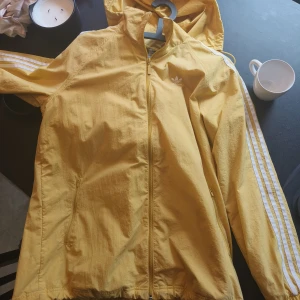 Gul vindjacka från Adidas med huva - Snygg gul vindjacka från Adidas med klassiska vita ränder längs ärmarna och vit logga på bröstet. Jackan har dragkedja framtill, två fickor och en praktisk huva. Perfekt för dig som gillar sportig och avslappnad stil.