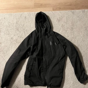Svart vindjacka från Helly Hansen - Svart vindjacka från Helly Hansen med huva och dragkedja framtill. Jackan har två sidofickor, en bröstficka med dragkedja och diskret HH-logga på bröstet. Tillverkad i lätt syntetmaterial med meshfoder för extra komfort. Perfekt för blåsiga dagar.