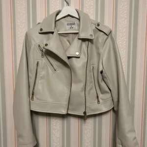 Beige skinnjacka från Stories S - Snygg beige skinnjacka från Stories i storlek S. Jackan är croppad med coola dragkedjor, axelklaffar och klassisk bikerkrage. Perfekt för dig som vill ha en stilren men edgy look. Materialet är mjukt och jackan har flera fickor med dragkedja.