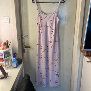 Klänning  - Superfin långklänning i ljuslila med tunna axelband och blommigt mönster i rött, vitt och gult. Klänningen har ett väldigt skönt material och är perfekt för varma dagar. Materialet känns mjukt och svalt mot huden.