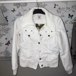 Vit jacka från Eve of Eden XS - Snygg vit jacka från Eve of Eden i storlek XS. Jackan har ribbade muddar och krage, två bröstfickor med knappar och coola detaljer framtill och baktill. Perfekt för dig som gillar en stilren look med lite edge.
