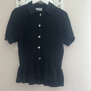 Svart kofta med volang från Holly & White S - Supersöt svart kofta från Holly & White med korta ärmar, volang nertill och ribbad struktur. Snygga pärlknappar framtill som ger en extra touch. Perfekt till jeans eller kjol för en trendig look.